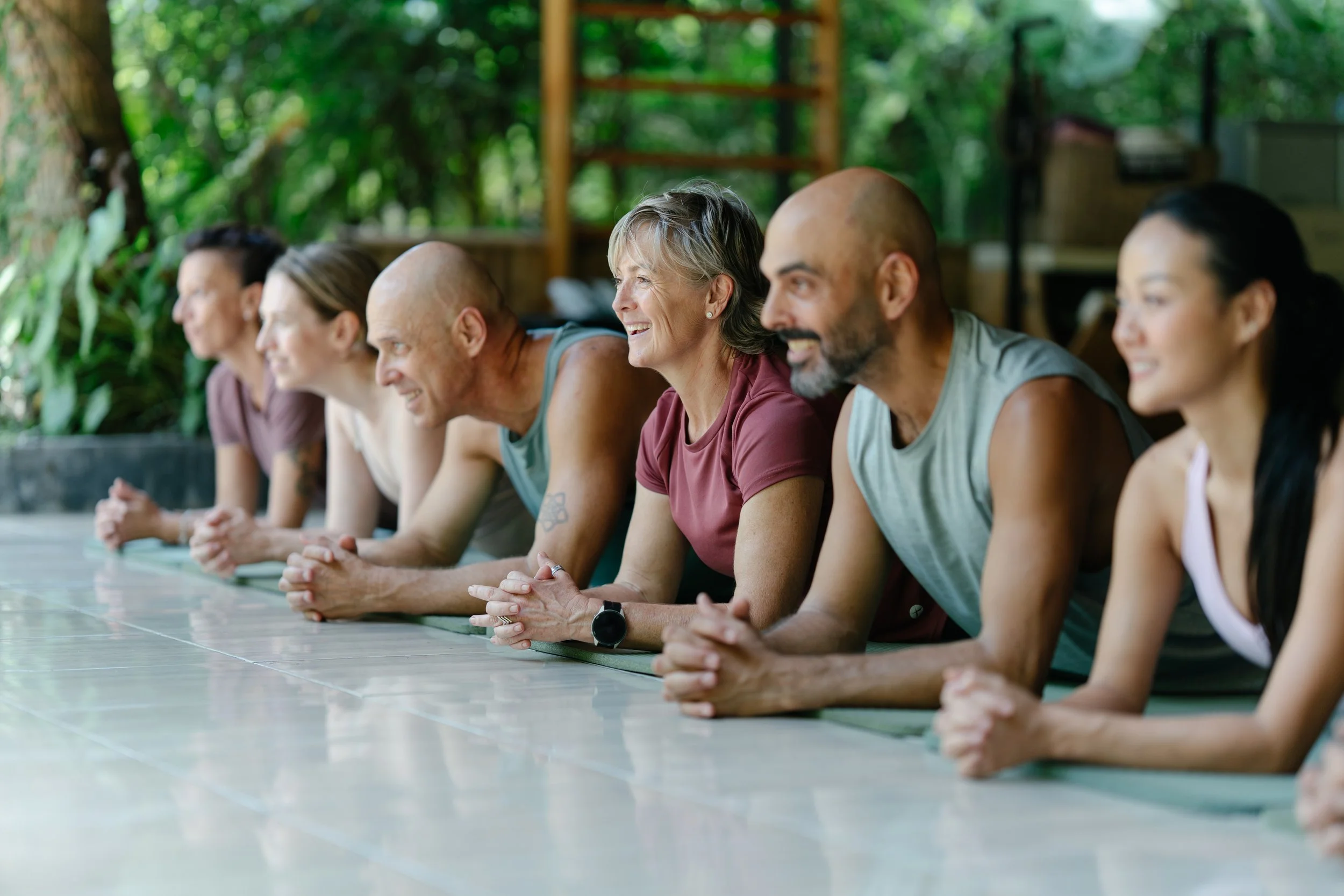 Group of people sitting on the floor in a yoga or meditation class outdoors, with greenery in the background.