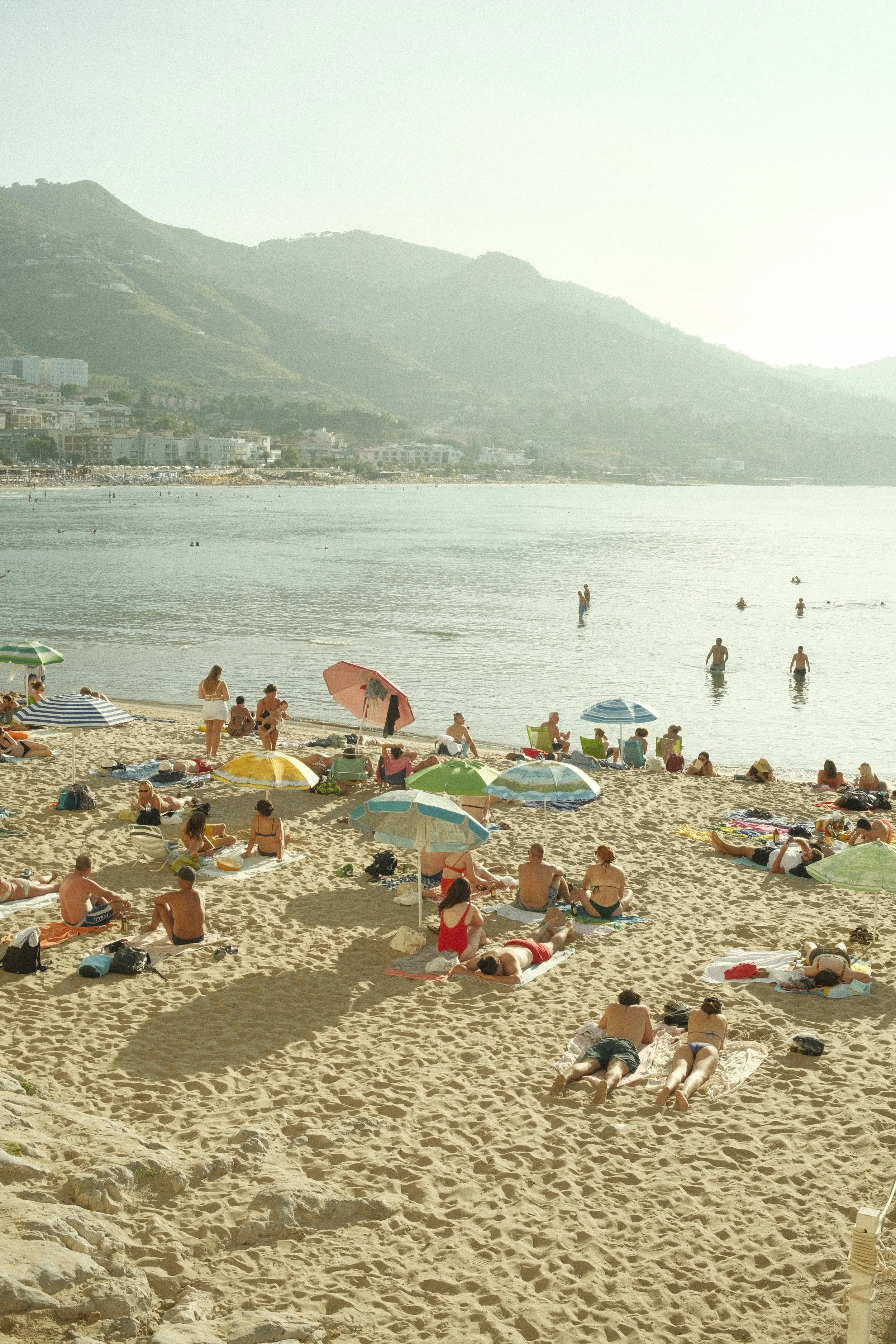Menschen entspannen am Strand, Sonnenliegen, Sonnenschirme, im Wasser schwimmen und sonnenbaden, im Hintergrund Berge und Gebäude, sonniges Wetter.