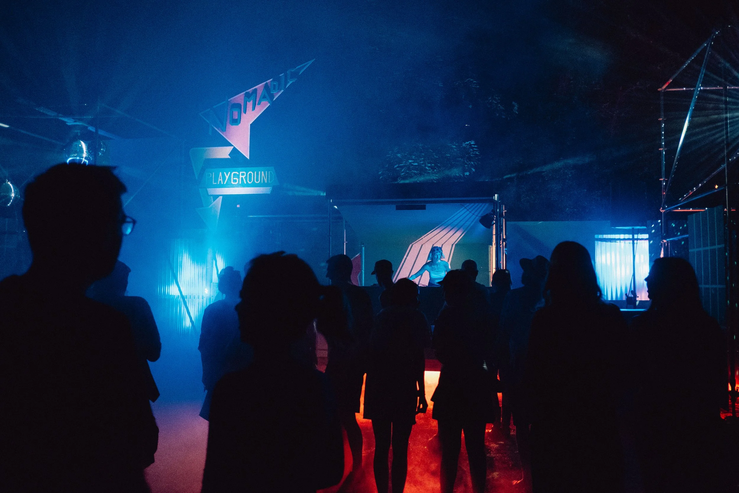 Menschen versammeln sich in einer dunklen, mit blauen und roten Lichtern beleuchteten Outdoor-Umgebung, bei der Bühne von Nomadic_Playground am Kesselfestival 2025.