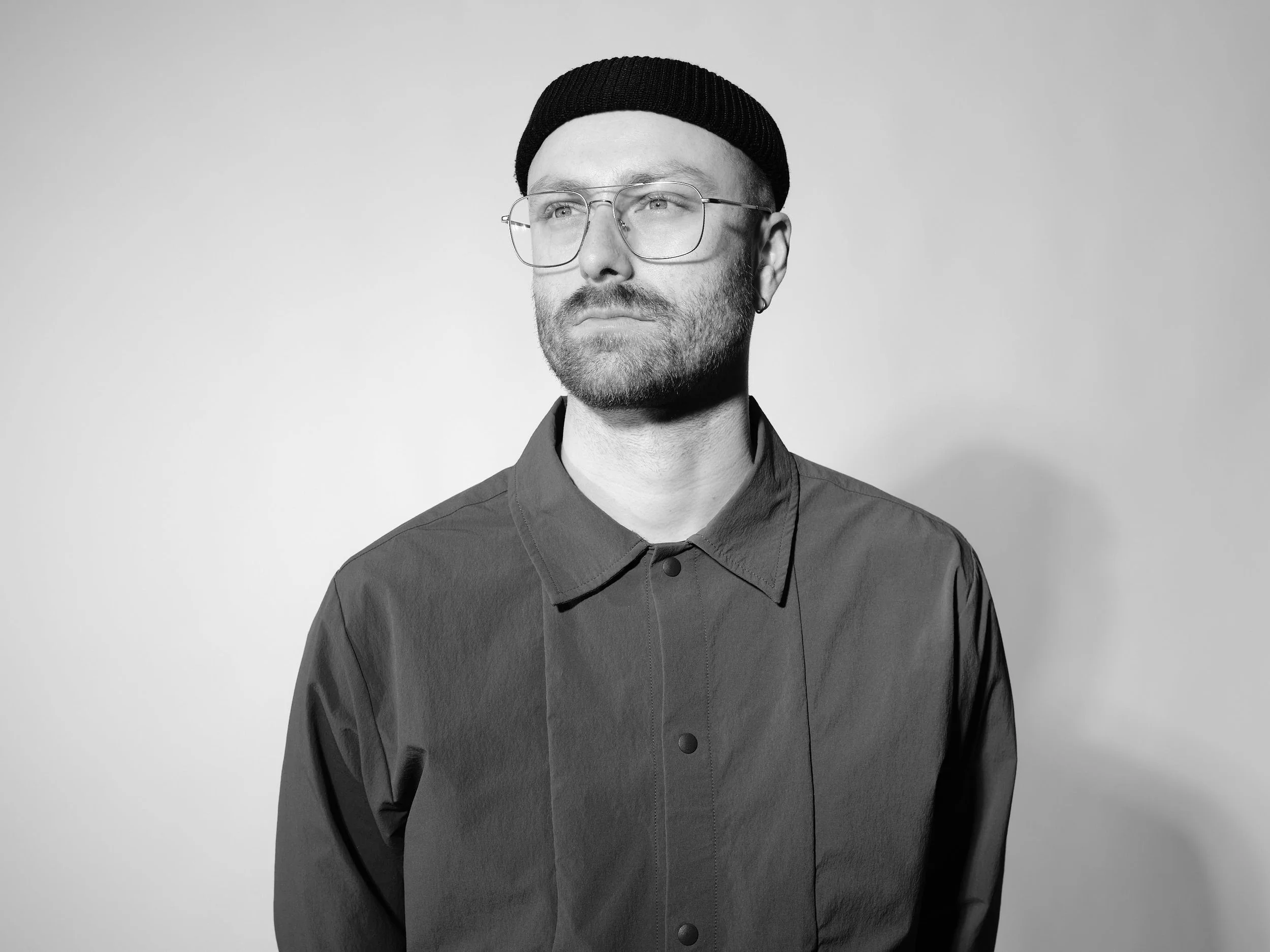 Schwarz-Weiß-Foto eines jungen Mannes mit Brille, schwarzem Beanie-Hut, dunklem Hemd und Earring. Blick nach rechts, neutraler Gesichtsausdruck, Kopf leicht nach links geneigt, Hintergrund grau. Inhaber von Kay Sigl Design