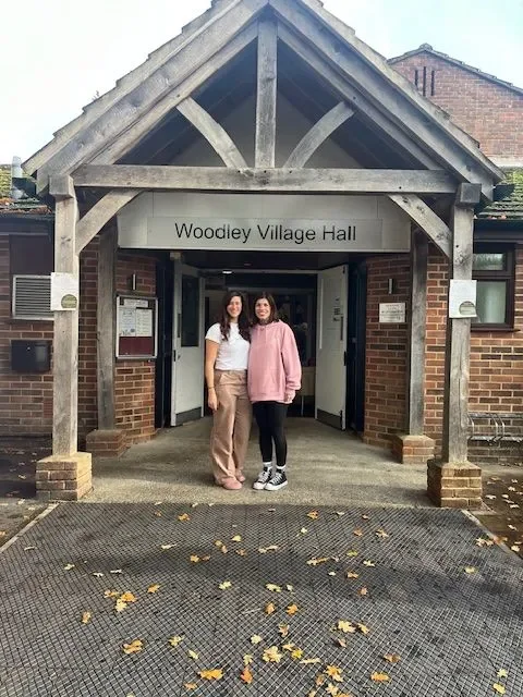 Two women standing under a wooden porch with a sign that reads 'Woodley Village Hall' on a brick building.