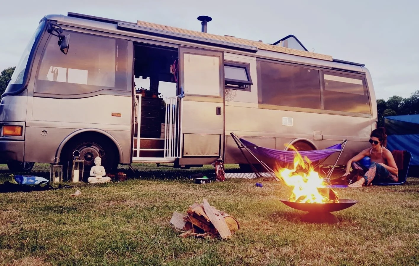A woman sitting outside a vintage campervan near a campfire at dusk.