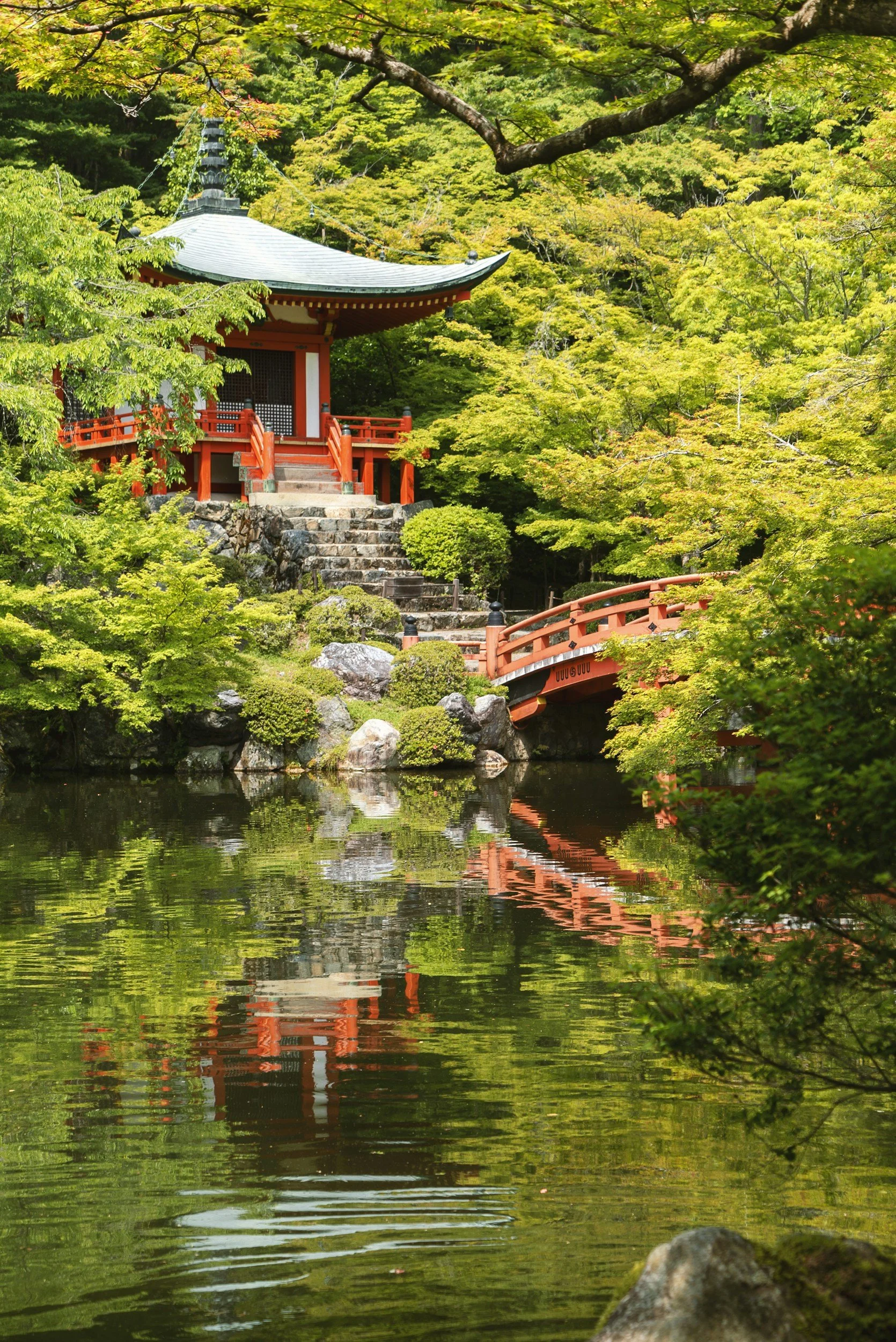 Traditional Japanese garden with a red and black wooden pavilion, a red arched bridge, lush green trees, and a pond reflecting the scenery.