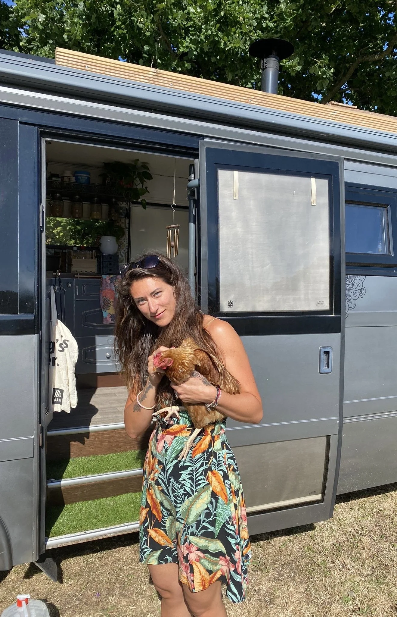 A woman with long brown hair holding a chicken outside of a silver and dark gray tiny house on wheels, with greenery and a tree in the background.