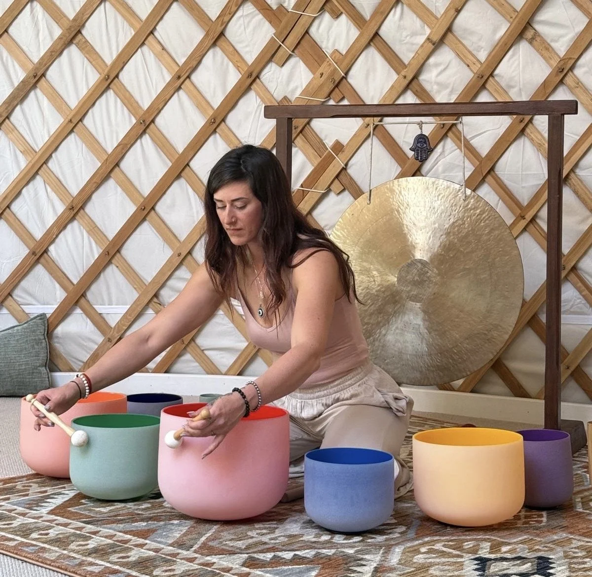 A woman sitting on a rug, playing a colorful crystal singing bowl set, with a large cymbal hanging on a stand behind her.