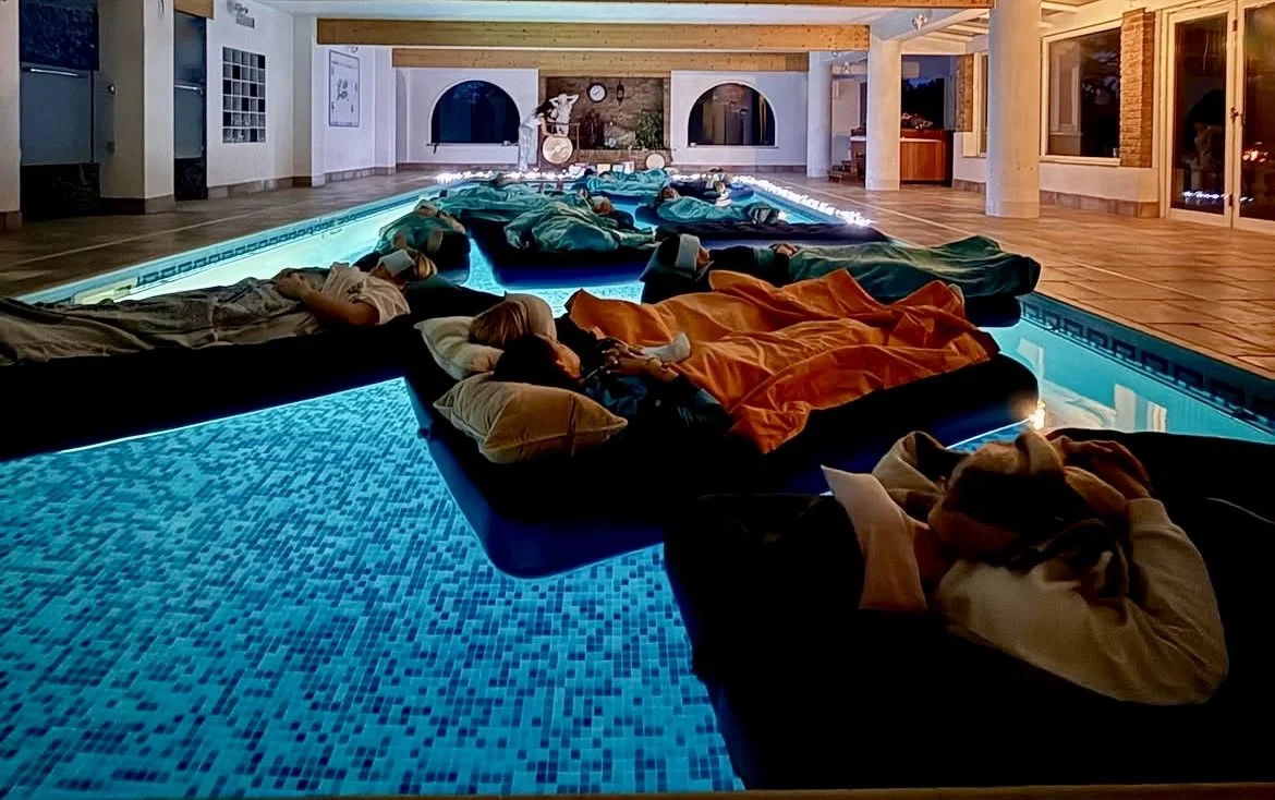 People sleeping on poolside lounge chairs with blankets and pillows inside a warmly lit indoor space with a swimming pool, large windows, and wooden beams.