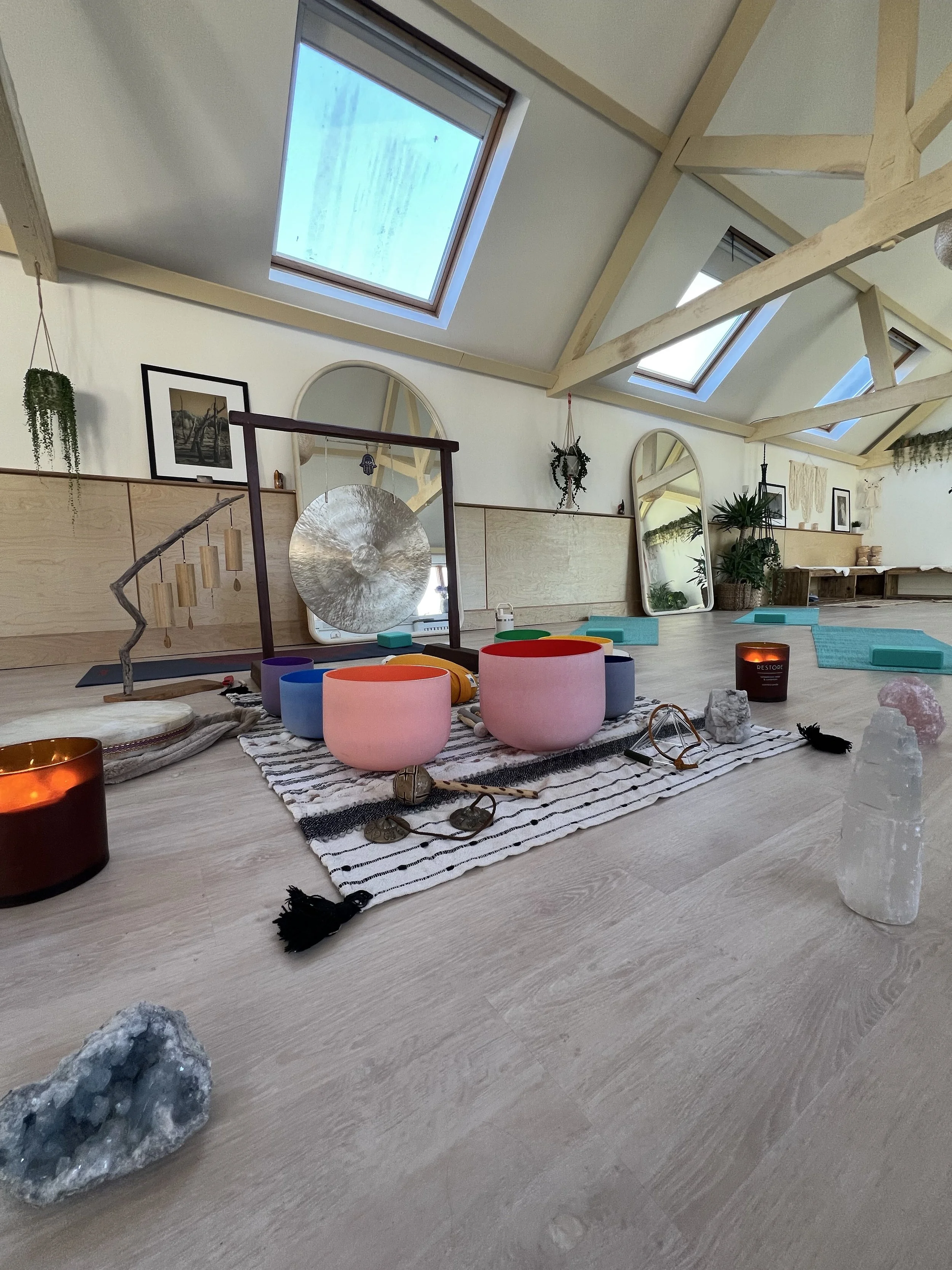 A peaceful yoga or meditation space in a bright room with skylight windows, wooden beams, mirrors, and plants. Various decorative objects, candles, and chimes are arranged on the floor.