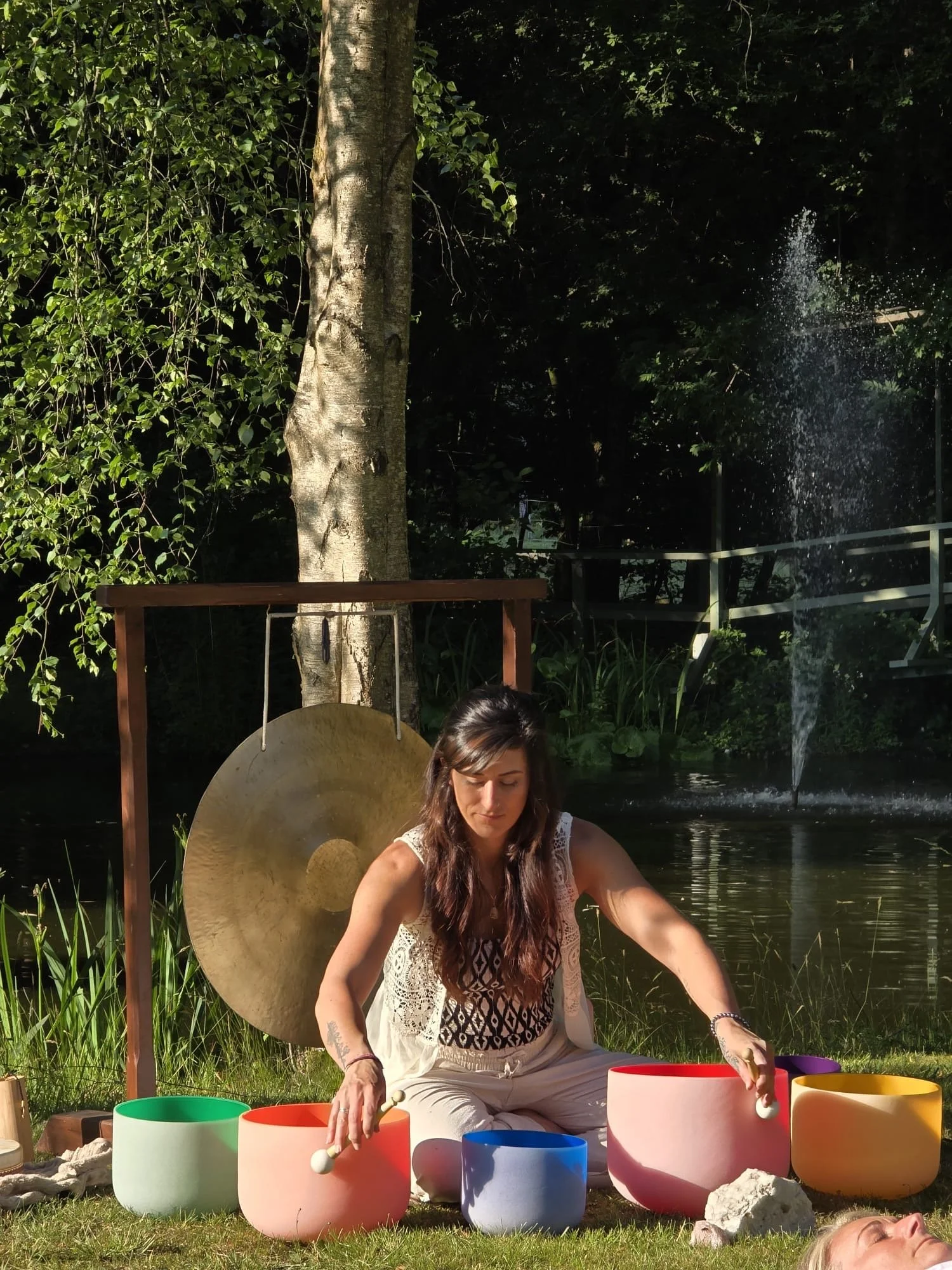 A woman playing crystal singing bowls outdoors near a pond and fountain, with trees and a gong nearby.