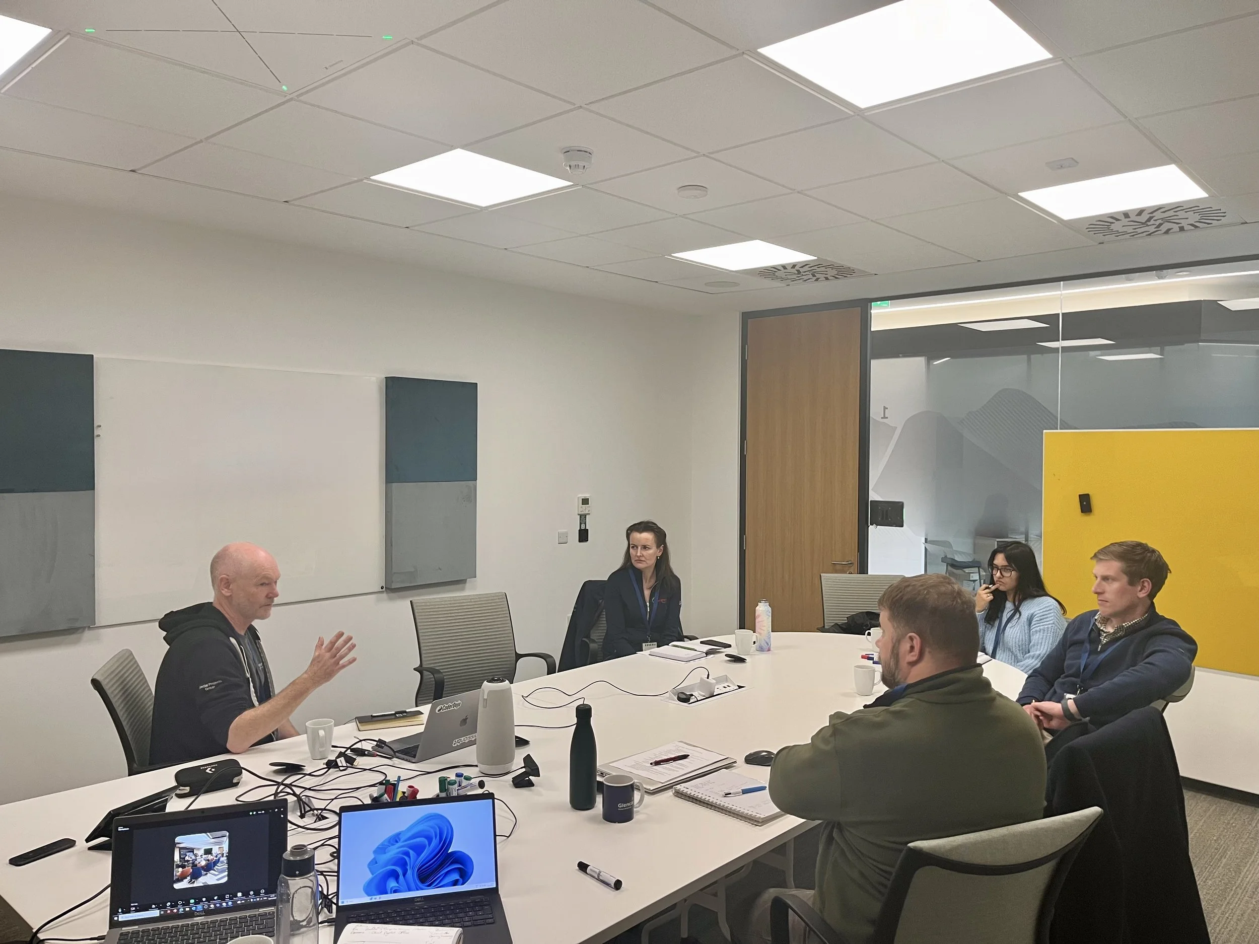 Five people attending a meeting in a modern conference room, seated at a large white table with laptops and notebooks, engaging in a discussion.