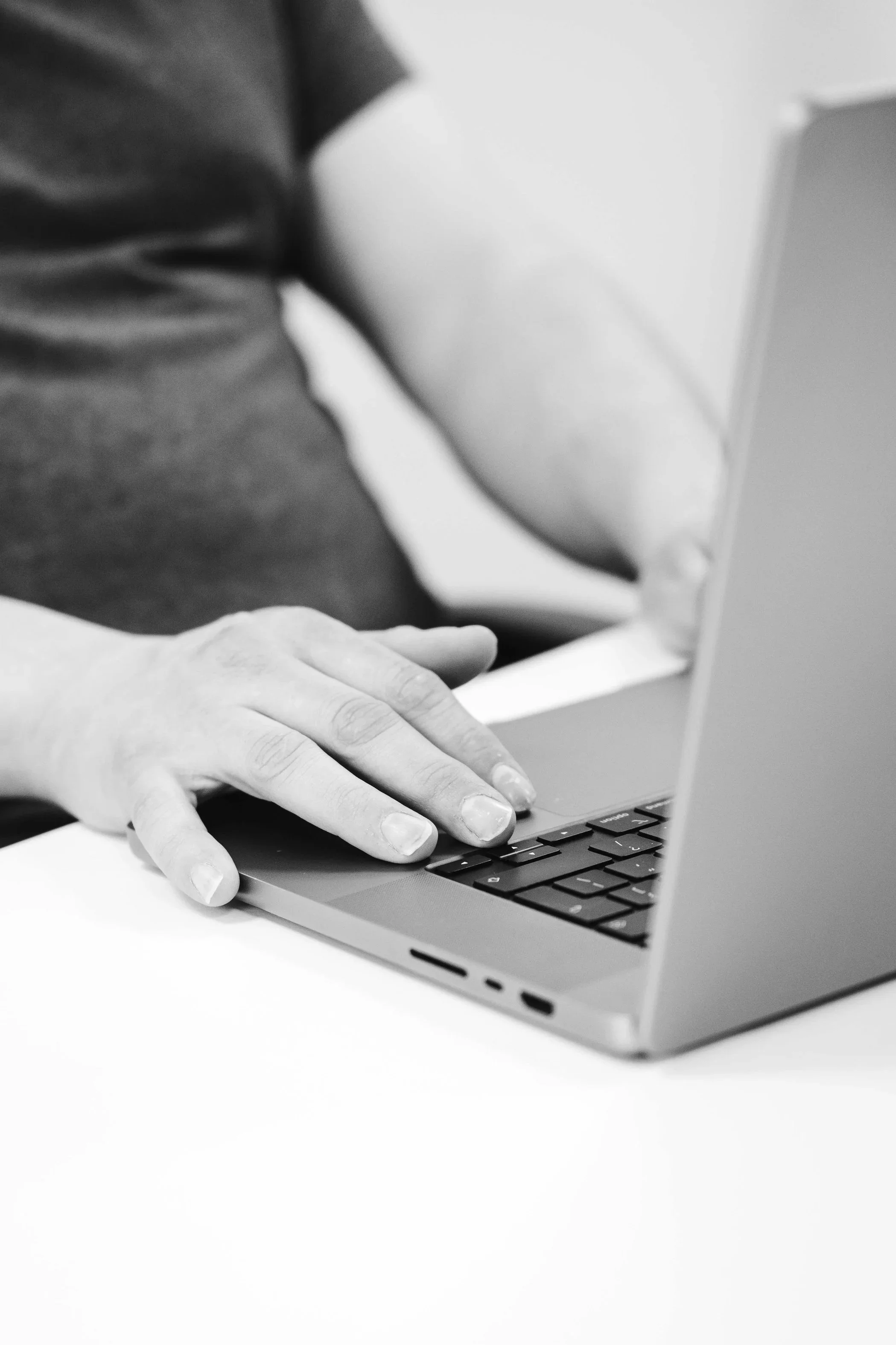 Close-up of a person’s hand typing on a laptop keyboard, with the person seated at a desk in an indoor setting.