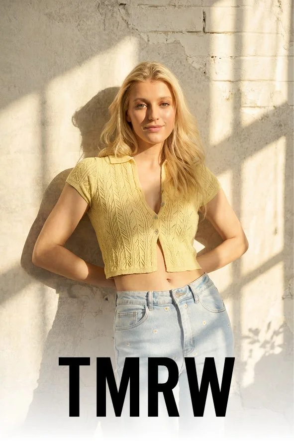 Junge Frau mit blonden Haaren in gelbem Cardigan und Jeans lehnt an einer hellen Wand mit Schatten im Sonnenlicht.