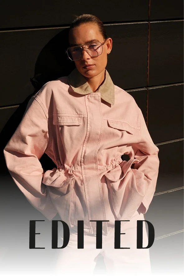 Junge Frau trägt eine rosa Jacke mit beige Kragen, steht vor einer schwarzen Wand und schaut ernst in die Kamera, Sonnenlicht wirft Schatten. Das Wort 'EDITED' steht unten im Bild.