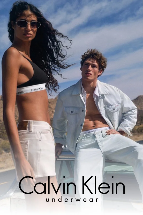 Ein Mann und eine Frau posieren im Freien bei sonnigem Wetter, tragen weiße Calvin Klein Unterwäsche und eine Jacke, umgeben von einer Wüstenlandschaft.