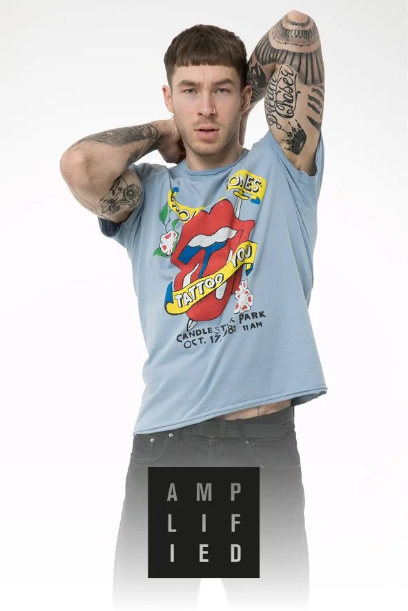 Junge Mann mit Tattoos trägt ein graues T-Shirt mit einem Rolling Stones Zungen-Logo und Text, posiert gegen weißen Hintergrund.
