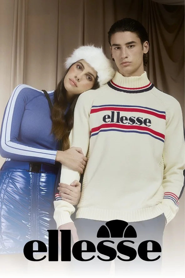 Ein junges Paar steht vor einem neutralen Hintergrund, das Mädchen trägt einen blauen Oberteil mit weißen Streifen an den Ärmeln und eine weiße Fellmütze, während der Junge ein cremefarbenes sweater mit 'ellesse'-Markierung trägt.