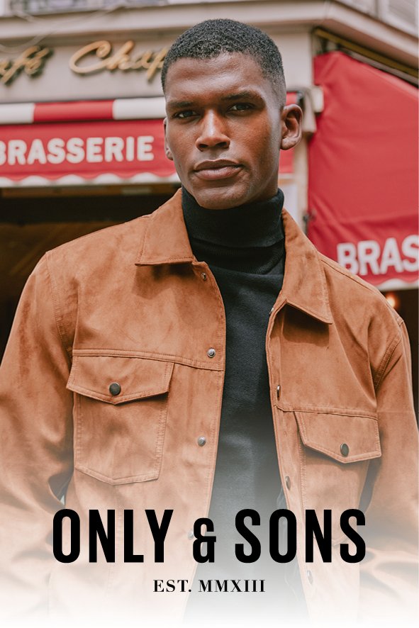 Ein junger Mann mit kurzen Haaren trägt eine braune Jacke und einen schwarzen Rollkragenpullover, steht vor einer brasserie mit roter Markise in Paris.