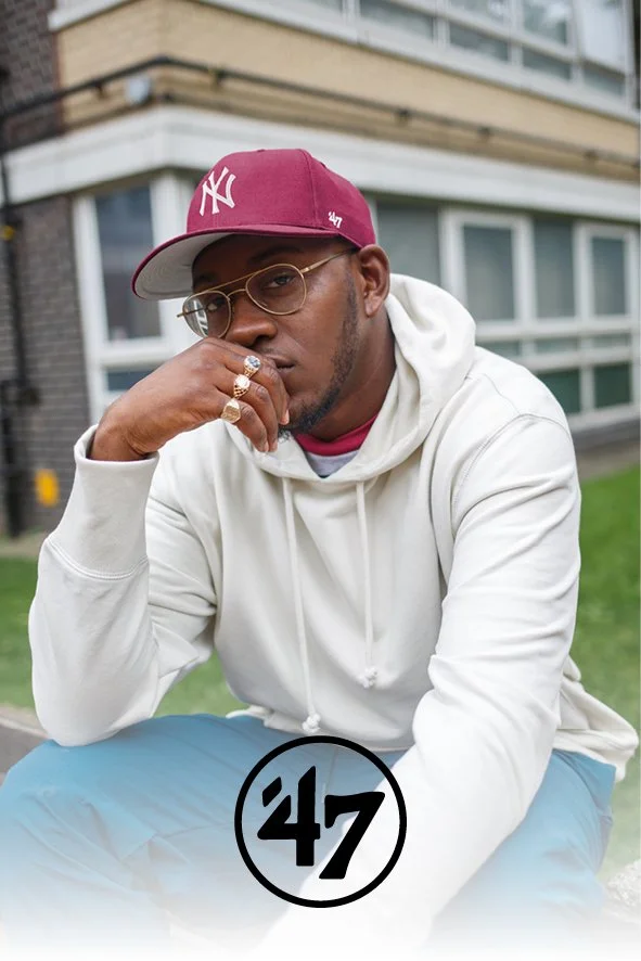 Ein Mann sitzt im Freien vor einem modernen Gebäude, trägt eine rote Kappe mit 'NY' Logo, eine Brille, einen weißen Hoodie, blaue Hosen und mehrere Ringe. Auf dem Bild steht die Nummer 47.
