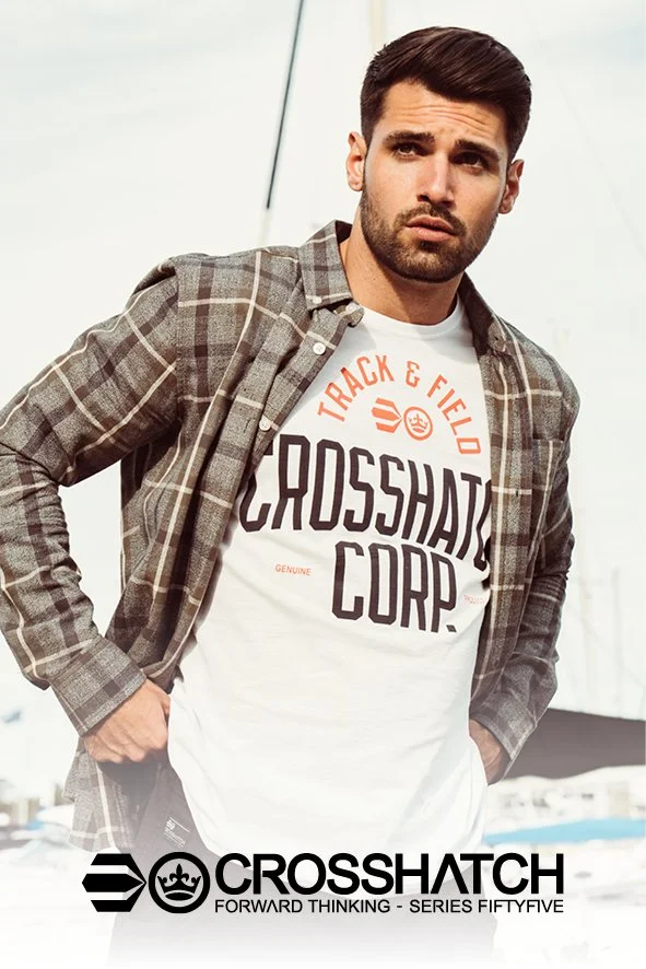 Mann mit dunklem Haar, Bart und gestreifter Jacke vor einem weißen Hintergrund, trägt ein weißes T-Shirt mit ‚Crosshatch‘-Schriftzug und logo, hält die Hände in den Taschen.