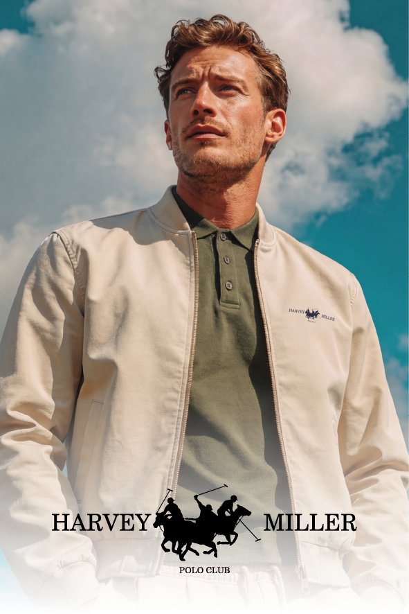 Mann mit braunen, lockigen Haaren trägt eine weiße Jacke und ein grünes Polo-Shirt, im Hintergrund blauer Himmel mit Wolken.