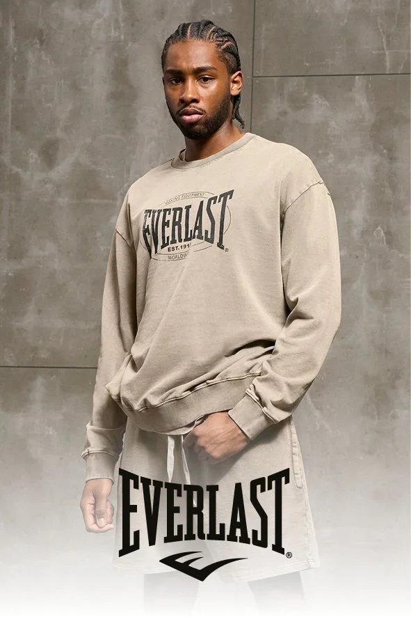 Ein jugendlicher Mann mit dunkler Hautfarbe und professionell gestylten Haaren steht vor einer grauen Betonwand. Er trägt ein beige Sweatshirt mit dem Logo "Everlast" und eine weiße Shorts mit dem gleichen Logo, das groß auf den Boden gedruckt ist.