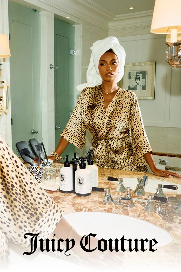 Eine Frau steht vor einem Spiegel im Badezimmer, trägt ein leopardmusterndes Kleid und ein Handtuch im Haar, umgeben von Pflegeprodukten.