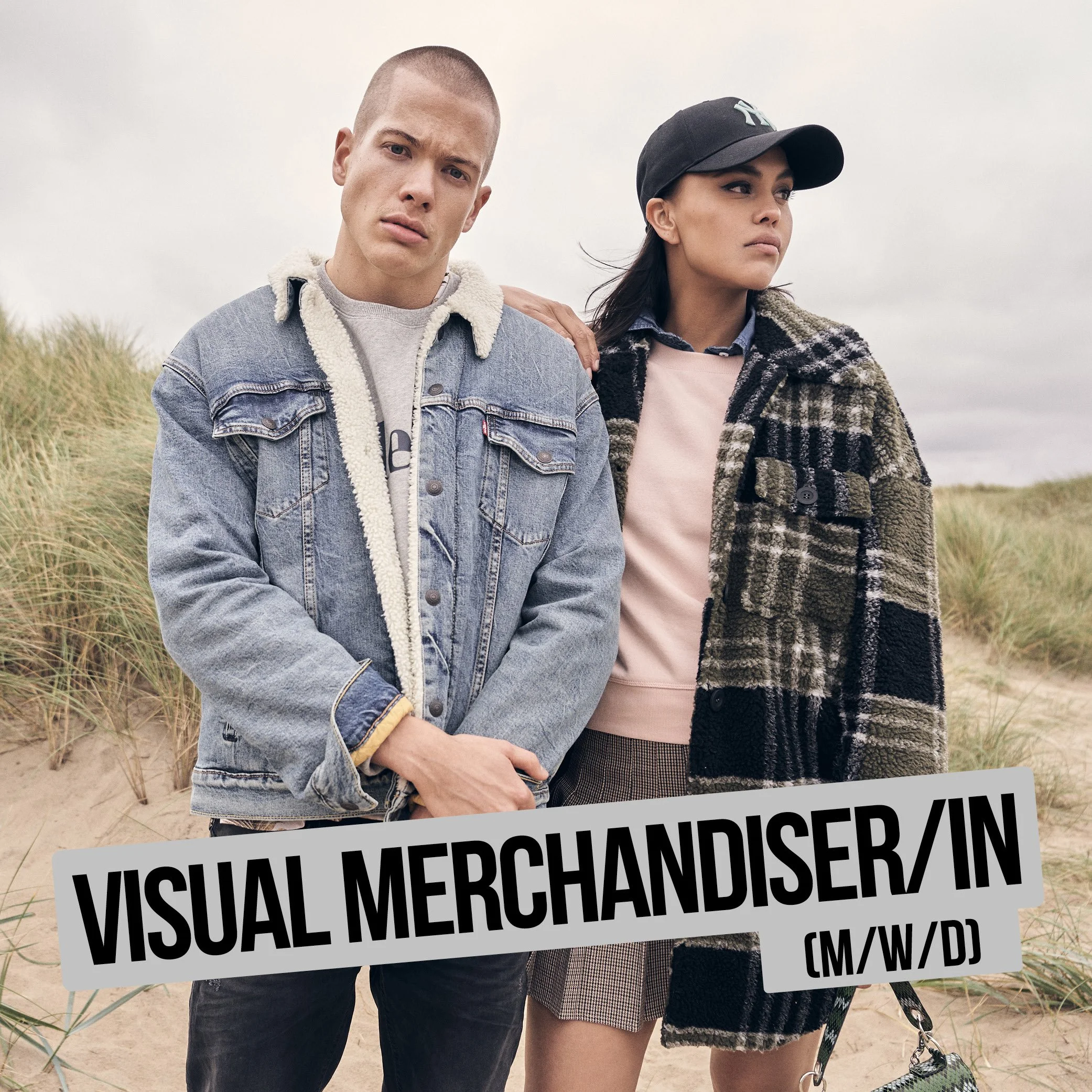 Zwei Menschen halten ein Schild mit der Aufschrift 'VISUAL MERCHANDISER' auf einem Strand. Der Mann trägt eine Jeansjacke mit Fellkragen, das Mädchen trägt eine Mütze und eine Jacke.