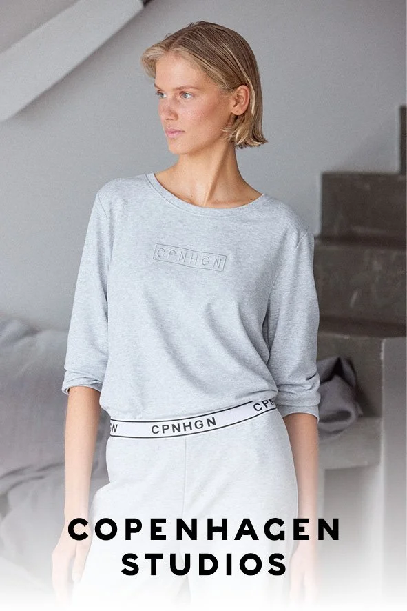 Eine junge Frau trägt ein graues Sweatshirt und eine passende Jogginghose in einem modernen, minimalistischen Raum.