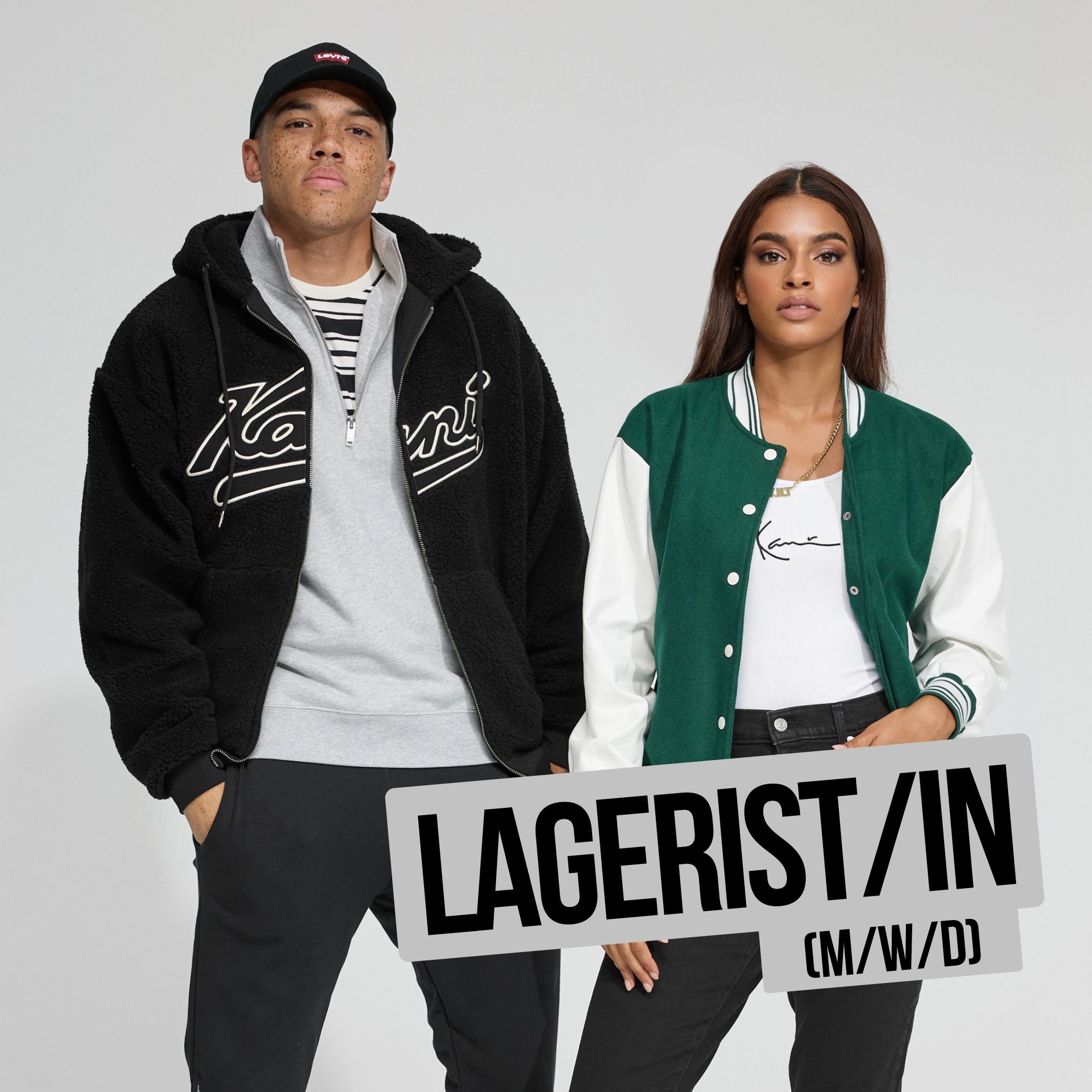 Zwei junge Menschen halten Plakate mit der Aufschrift ‚LASSERISMUS!‘, einer trägt eine schwarz-weiße Jacke und einen schwarzen Baseball cap, der andere trägt eine grüne Jacke, ein weißes T-Shirt und schwarze Jeans, gegen einen grauen Hintergrund.