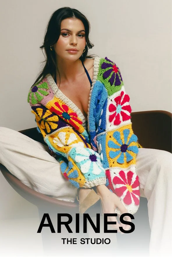 Eine junge Frau sitzt auf einem schwarzen Stuhl, trägt ein buntes, zuckergesteuertes Strickjäckchen mit großen Blumenmuster, beige Hosen, und blickt in die Kamera.