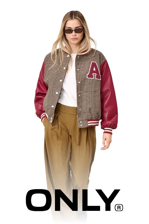 Junge Frau trägt eine Jacke mit roten Ärmeln, weißen Streifen an den Bündchen, und einem großen roten Buchstaben 'A' auf der Brust. Sie trägt Sonnenbrillen, ein weißes T-Shirt und braune, weite Hose.