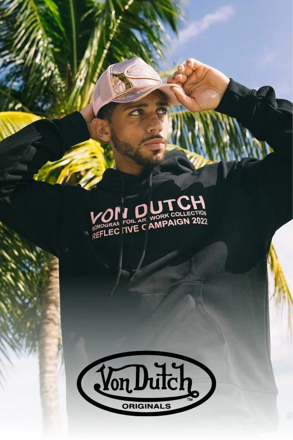 Ein junger Mann trägt einen schwarzen Hoodie mit dem Aufdruck 'Von Dutch Monogram Foil Air Work Collection Reflective Campaign 2022' und eine pink-weiße Baseballcap. Im Hintergrund sind Palmen und ein blauer Himmel zu sehen.