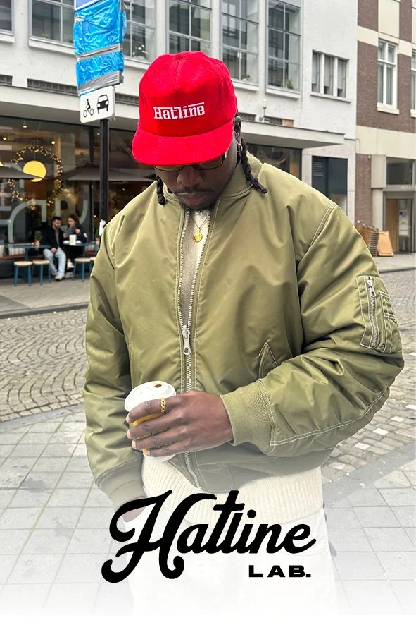 Ein Mann trägt eine rote Cap mit der Aufschrift 'Hatline' und eine grüne Bomberjacke, hält eine Tasse Kaffee in der Hand, steht auf einer Stadtstraße mit Cafés und Gebäuden im Hintergrund.