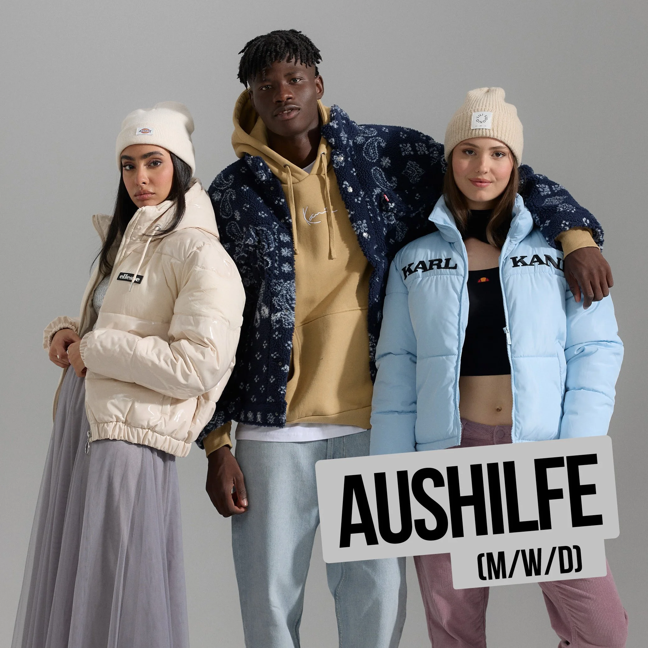 Drei junge Menschen in Streetwear mit Mützen posieren vor neutralem Hintergrund, einer hält ein Schild mit Text.