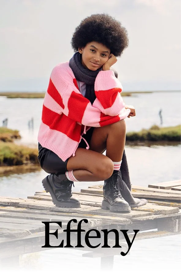 Junge Frau mit Afro-Haare, die auf einer Holzbrücke am Wasser sitzt, trägt eine pink-rote Strickjacke, schwarze Shorts, schwarze Stiefel und einen grauen Schal.