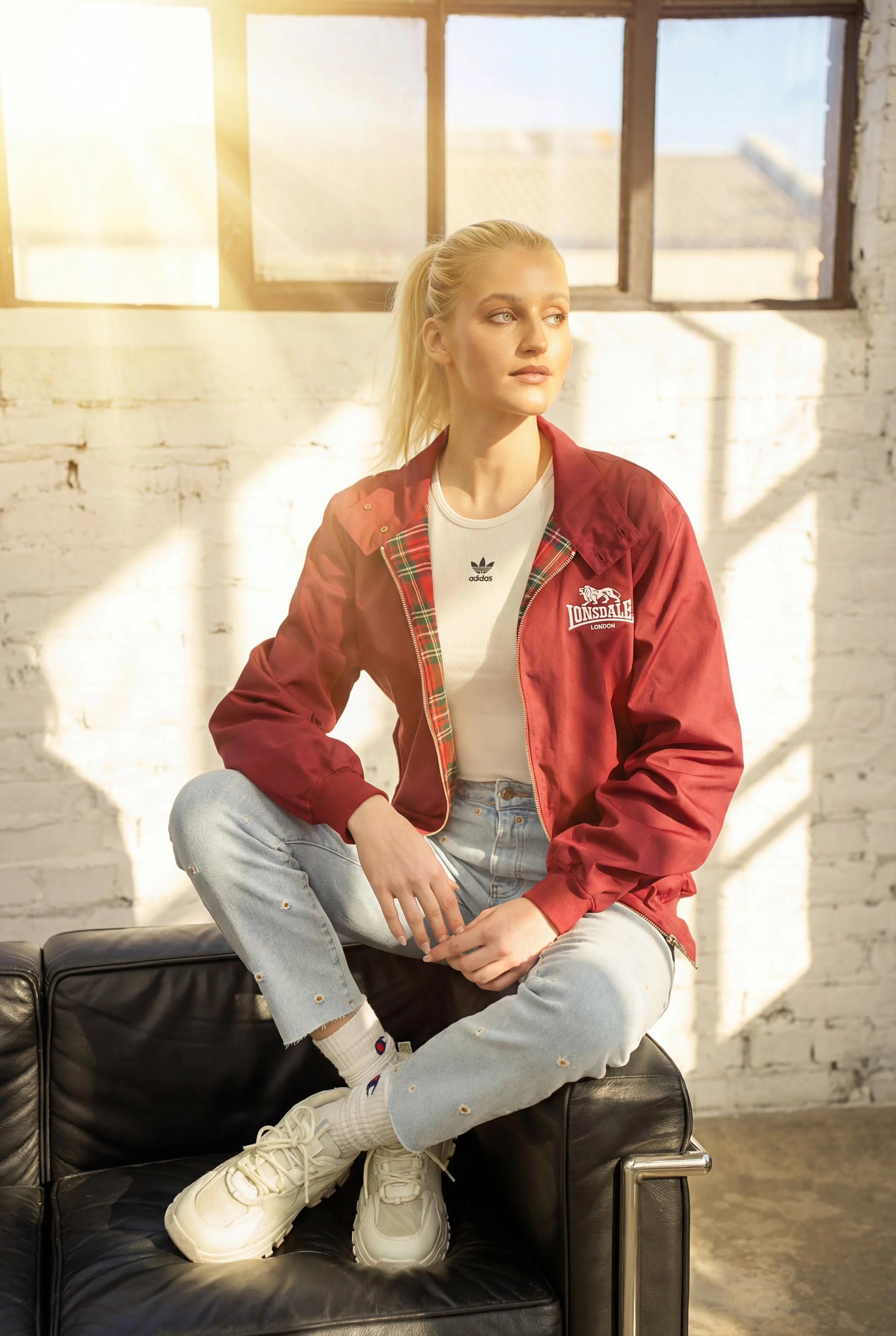 Junge Frau mit blonden Haaren sitzt auf einem schwarzen Sofa vor einer weißen Ziegelwand, trägt Jeans, weißen Turnschuhen, roten Lonsdale Jacke und blickt nach rechts.