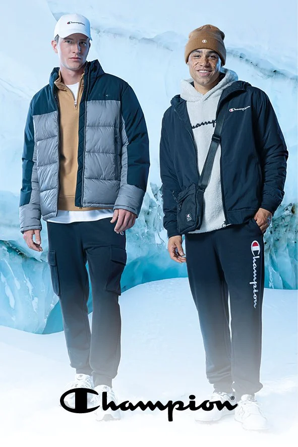 Zwei Männer in Winterkleidung stehen vor einer Eiswand in einer Snow- oder Eisszene, beide tragen Champion-Labels. Eines trägt eine beige Kapuzenjacke, das andere eine dunkle Jacke mit Schulter- und Ellenbogenaufsätzen. Sie tragen sportliche Hosen un