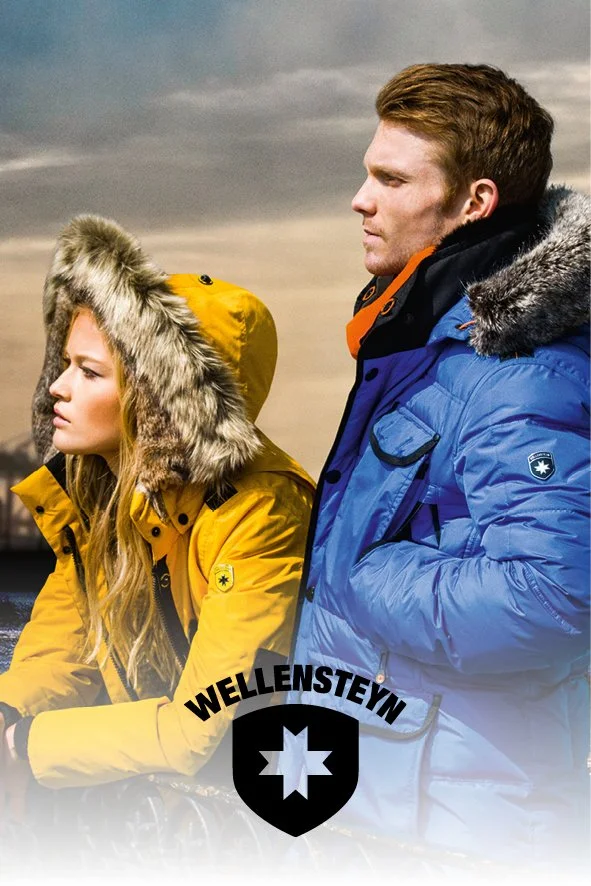 Ein Mann und eine Frau tragen Wintermäntel mit Fellkapuzen und schauen nachdenklich in die Ferne, im Hintergrund ein bewölkter Himmel, darunter das Wellenstein-Logo.