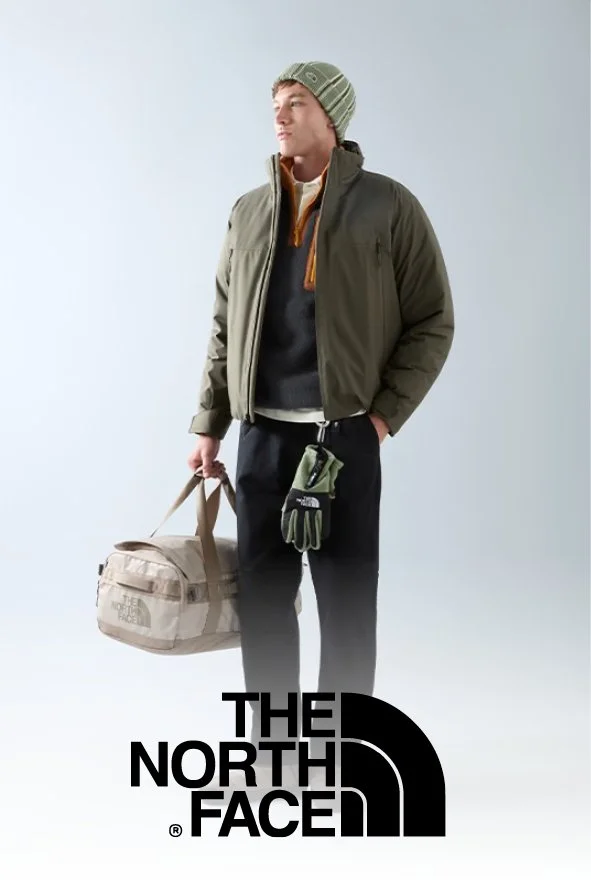 Ein junger Mann steht in Outdoor-Bekleidung vor einem hellen Hintergrund. Er trägt eine grüne Jacke, einen grau-grünen Wollmütze, eine schwarze Sweater mit Reißverschluss, schwarze Hosen und hält eine beige The North Face Tasche sowie Handschuhe in d