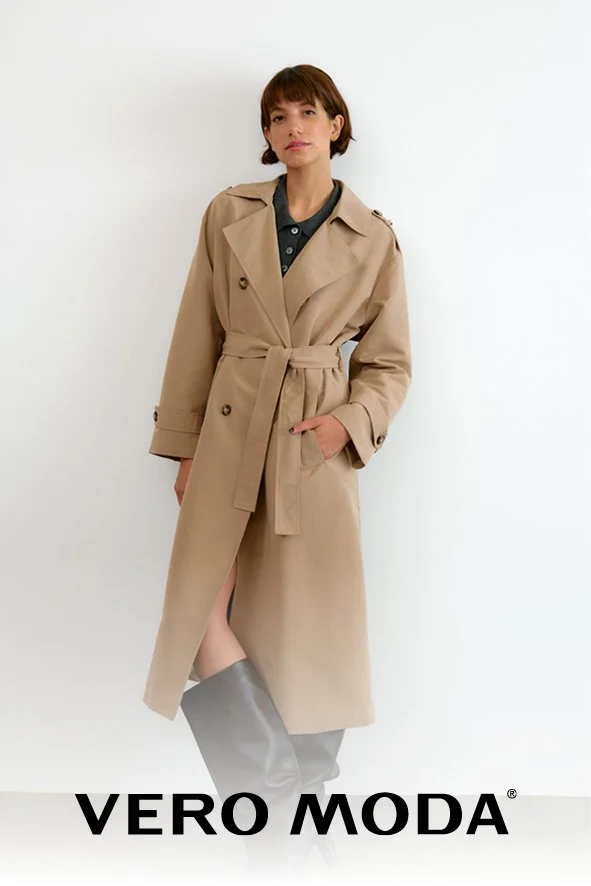 Frau in einem beige Mantel mit Gürtel, grauem Hemd und schwarzen Stiefeln steht vor weißem Hintergrund. Das Bild enthält den Text "VERO MODA" am unteren Rand.