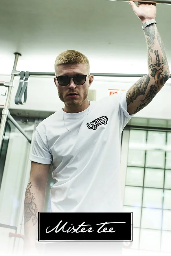 Ein junger Mann trägt eine weiße T-Shirt mit dem Logo "BarrelLong" und Sonnenbrille, steht in einer modernen, industrial Atmosphäre, vermutlich in einem Bus oder Zug, mit Tattoos an Armen und hält sich an einer Handstange fest. Unten befindet sich ei