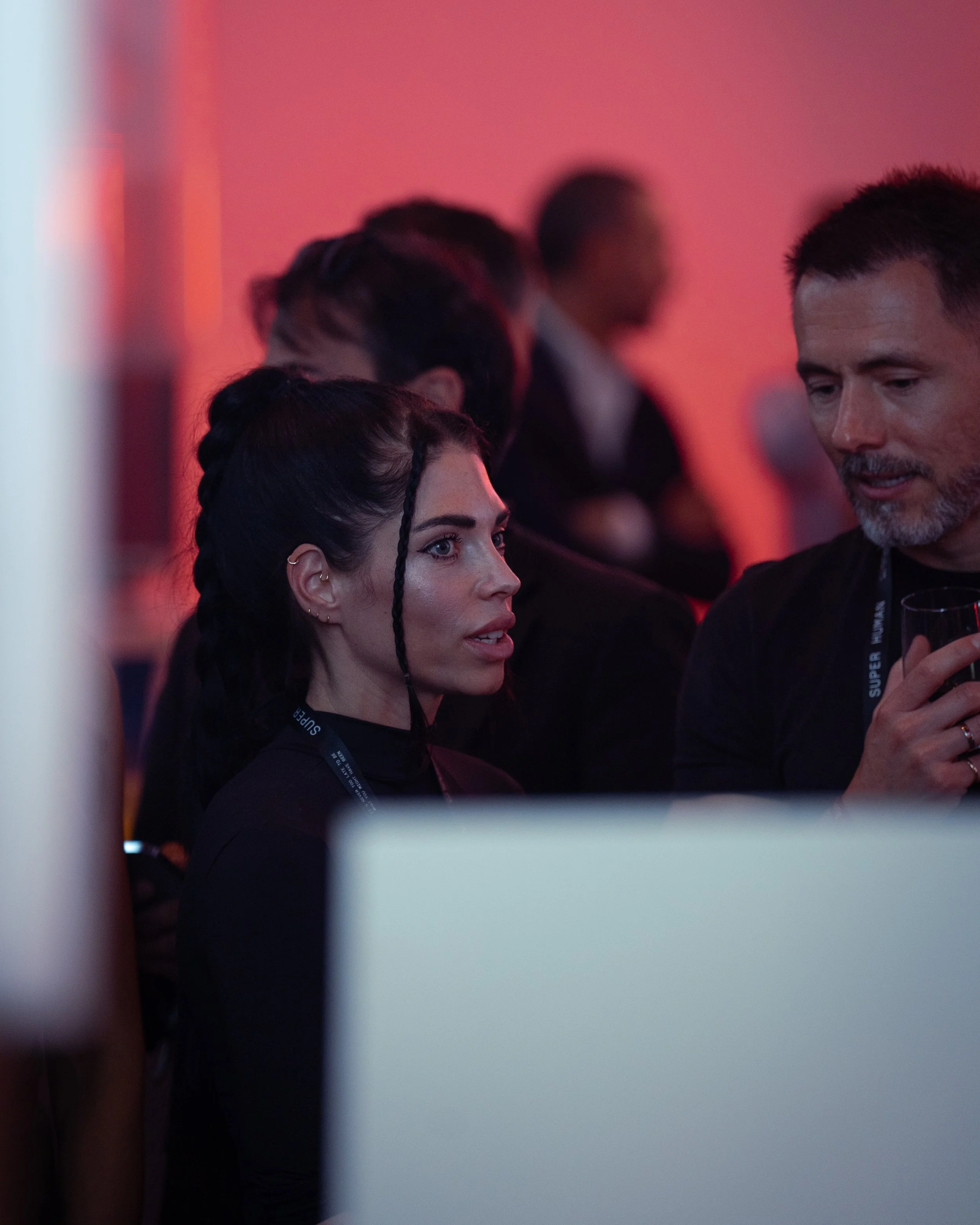 A woman with black hair styled in braids and wearing a black top is talking to a man holding a wine glass, in a dimly lit room with red lighting and blurred background of other people.