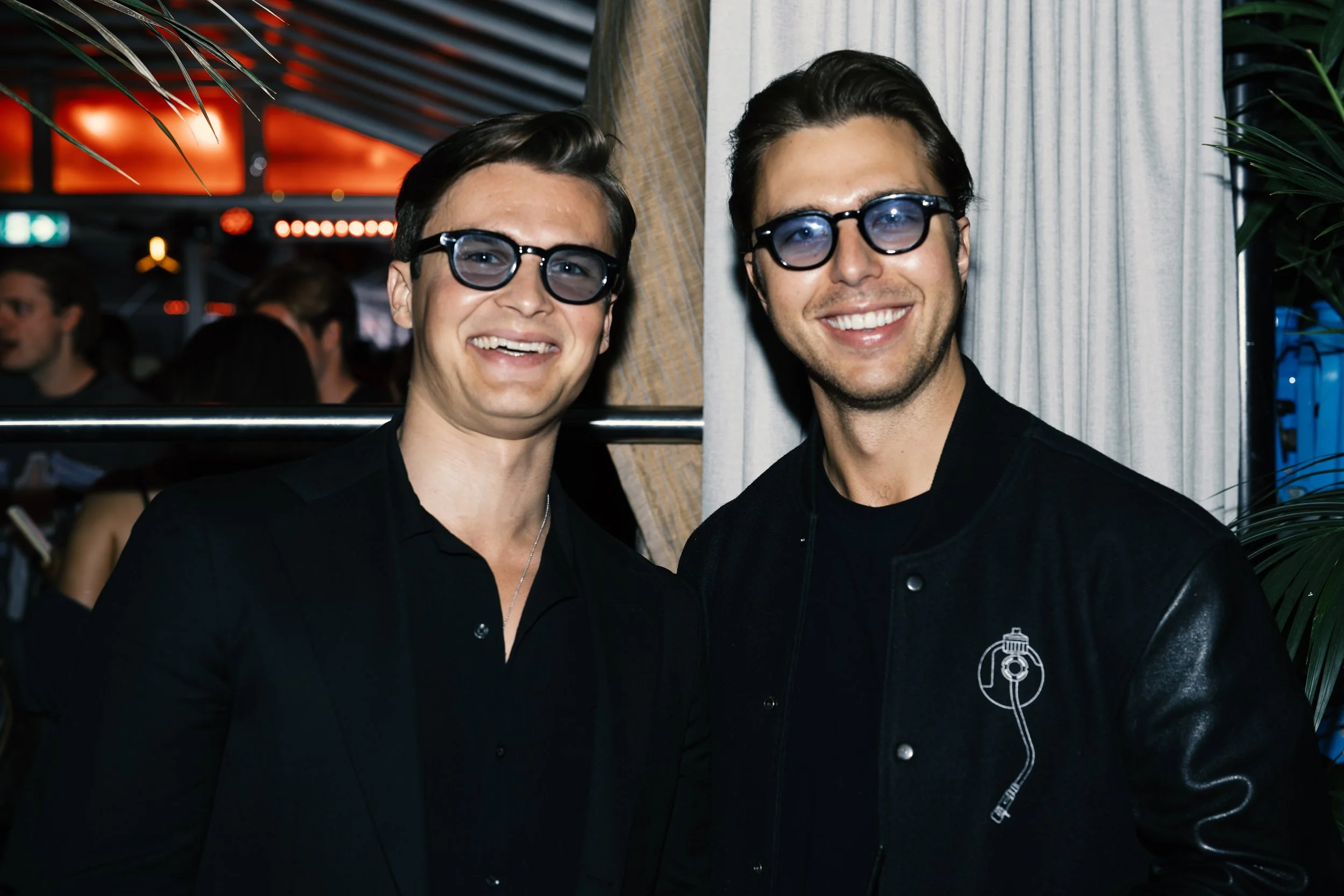 Two men wearing glasses and black clothing, smiling at the camera at a social event.
