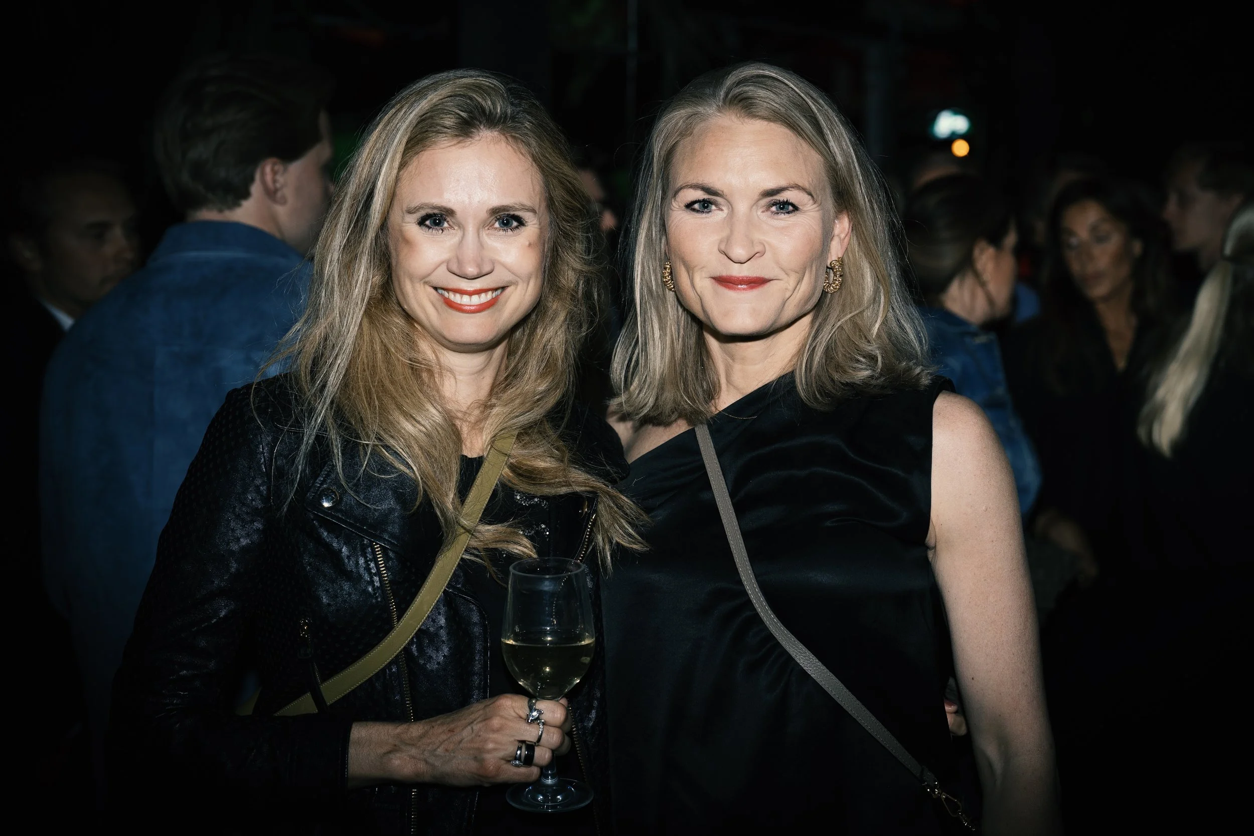 Two women smiling at a social event, one holding a glass of white wine, with a crowd of people in the background.