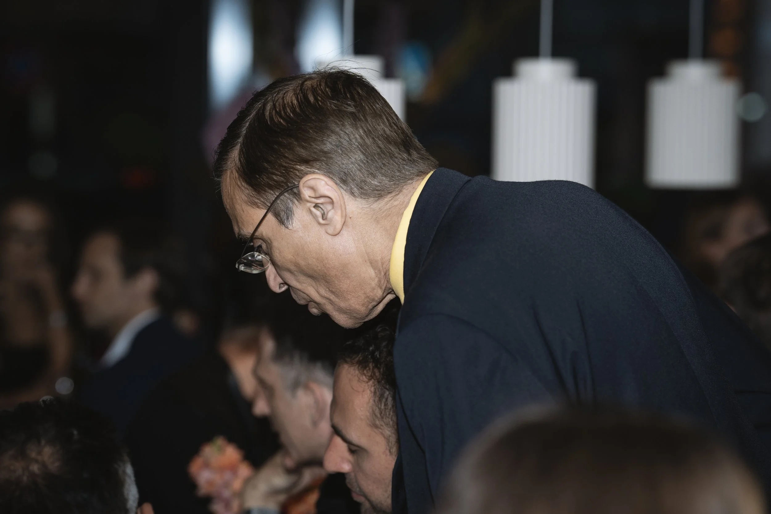 A man with glasses leaning over a seated man in a crowded indoor setting with soft lighting.