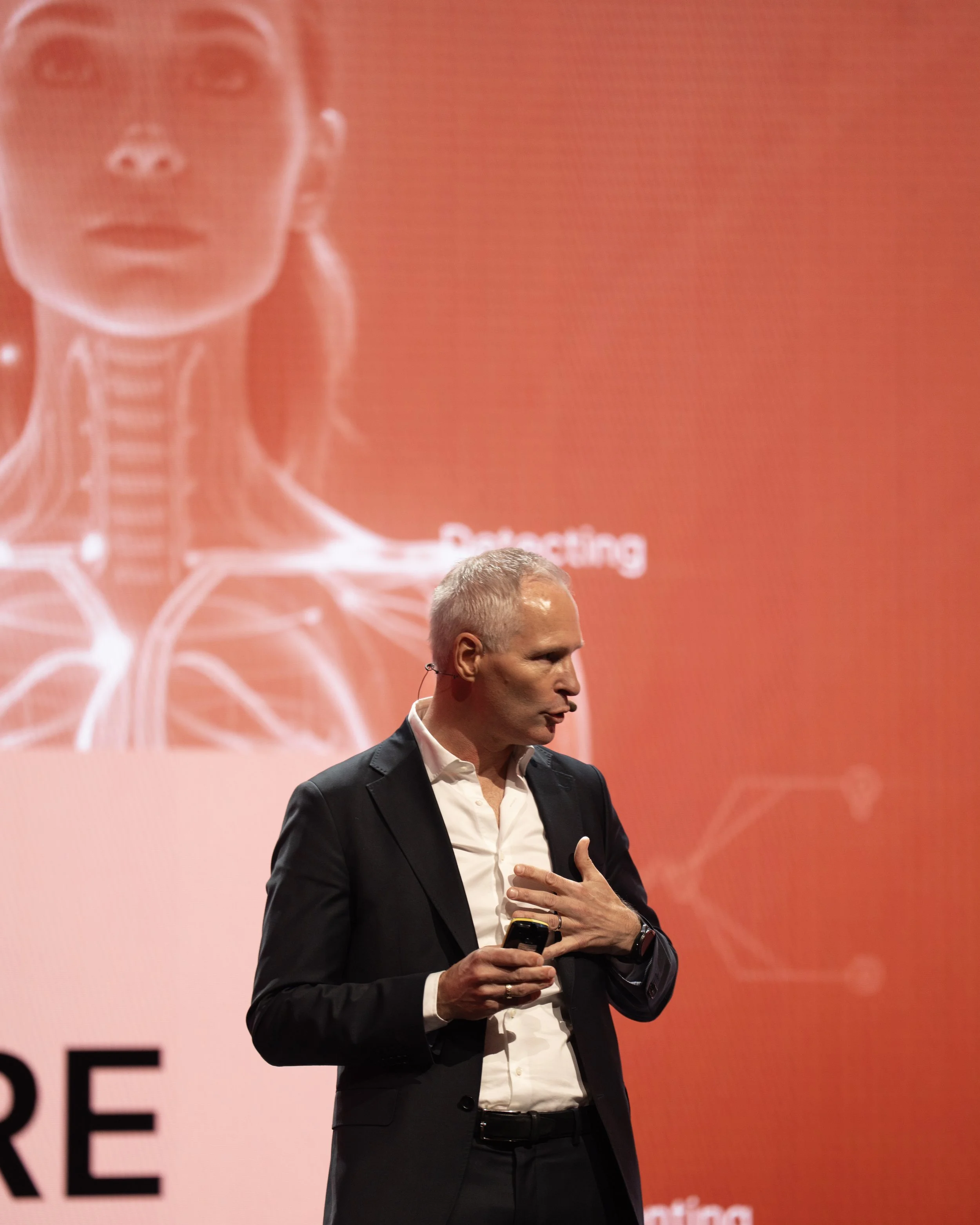A man in a black suit and white shirt giving a presentation on stage with a large red background and medical diagram of a human neck and thyroid gland.