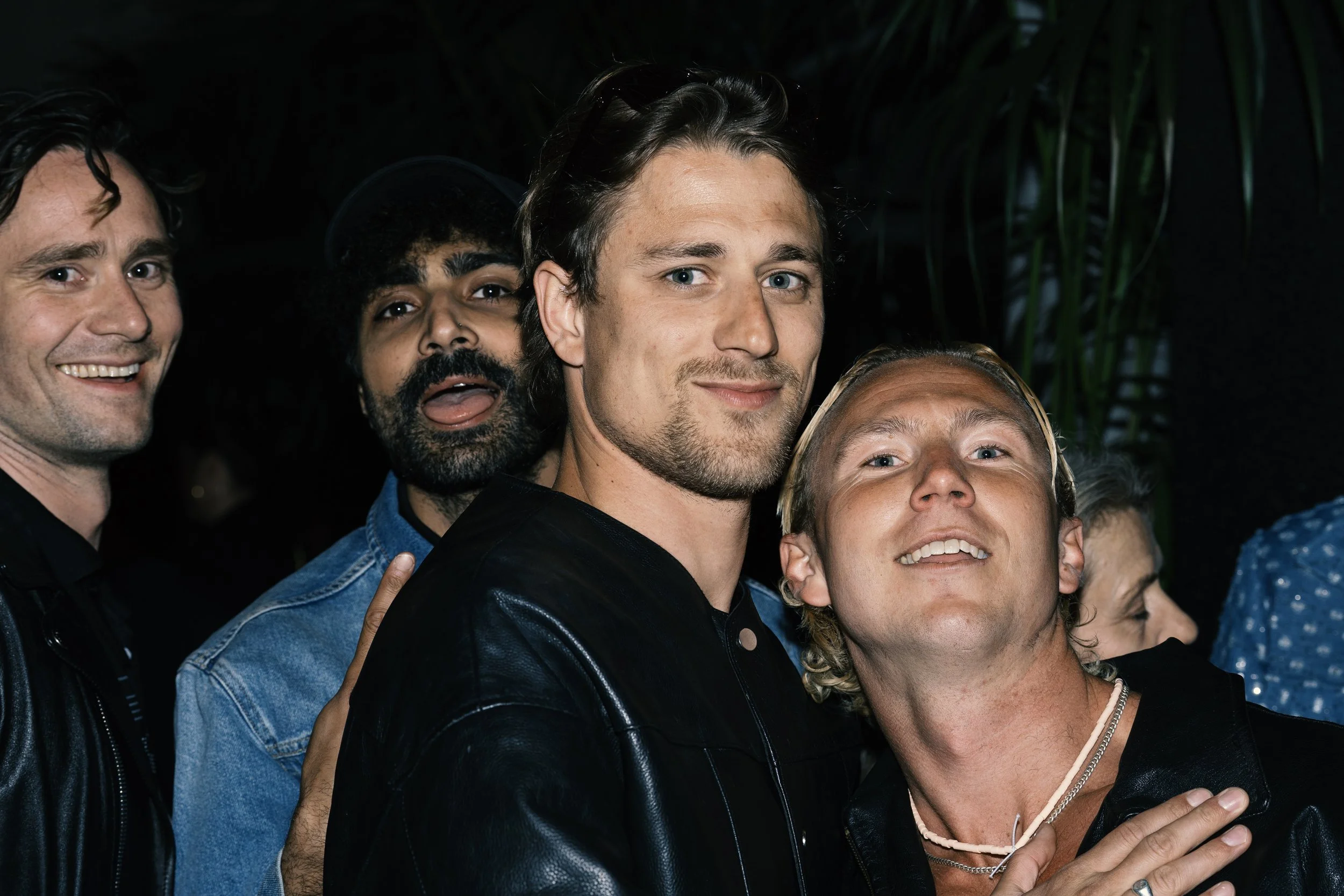 Group of five men taking a selfie together, smiling, in a dark environment with green plants in the background.