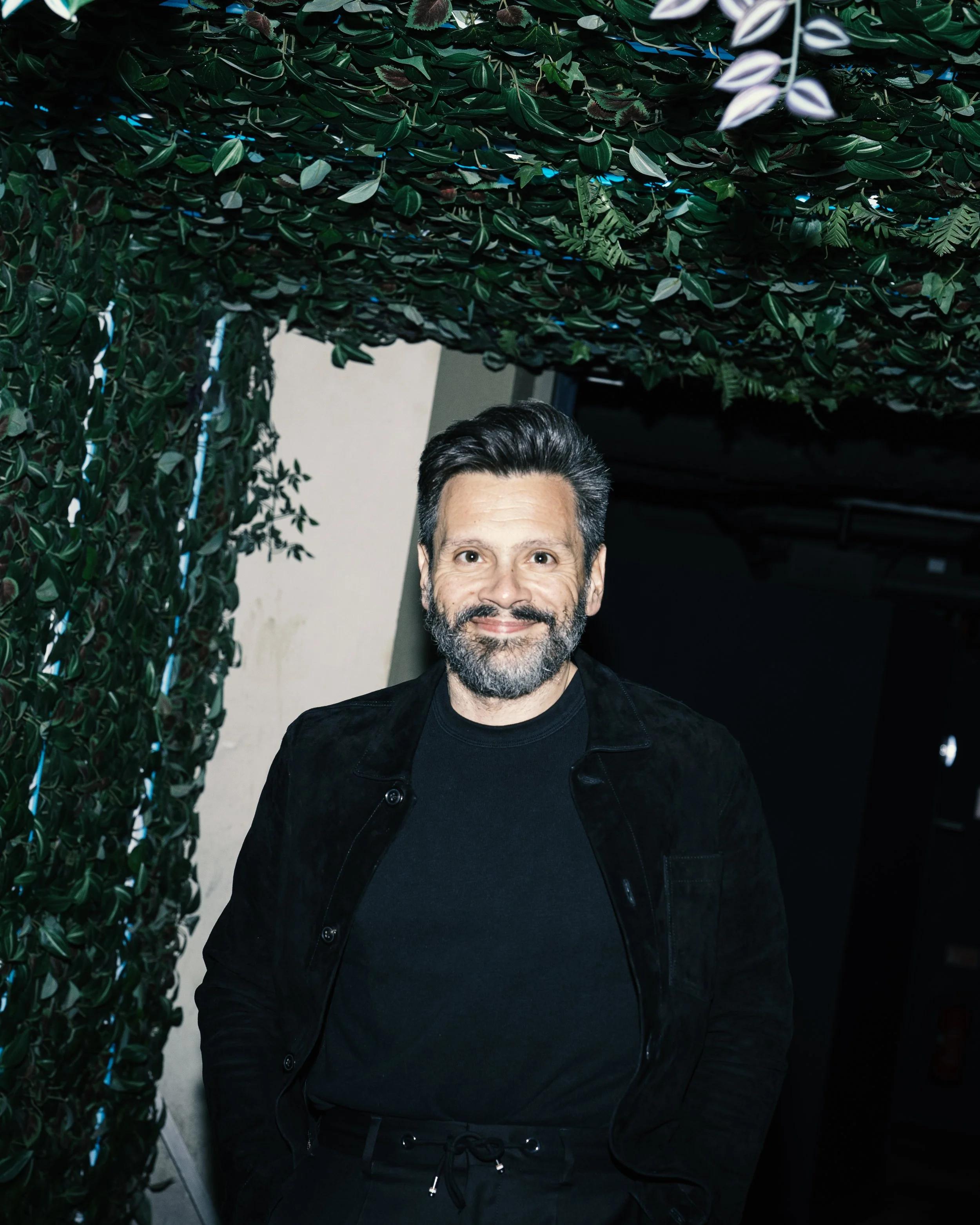 A man with dark hair and a beard smiling, wearing a black shirt and black jacket, standing under a decorated canopy of green leaves.