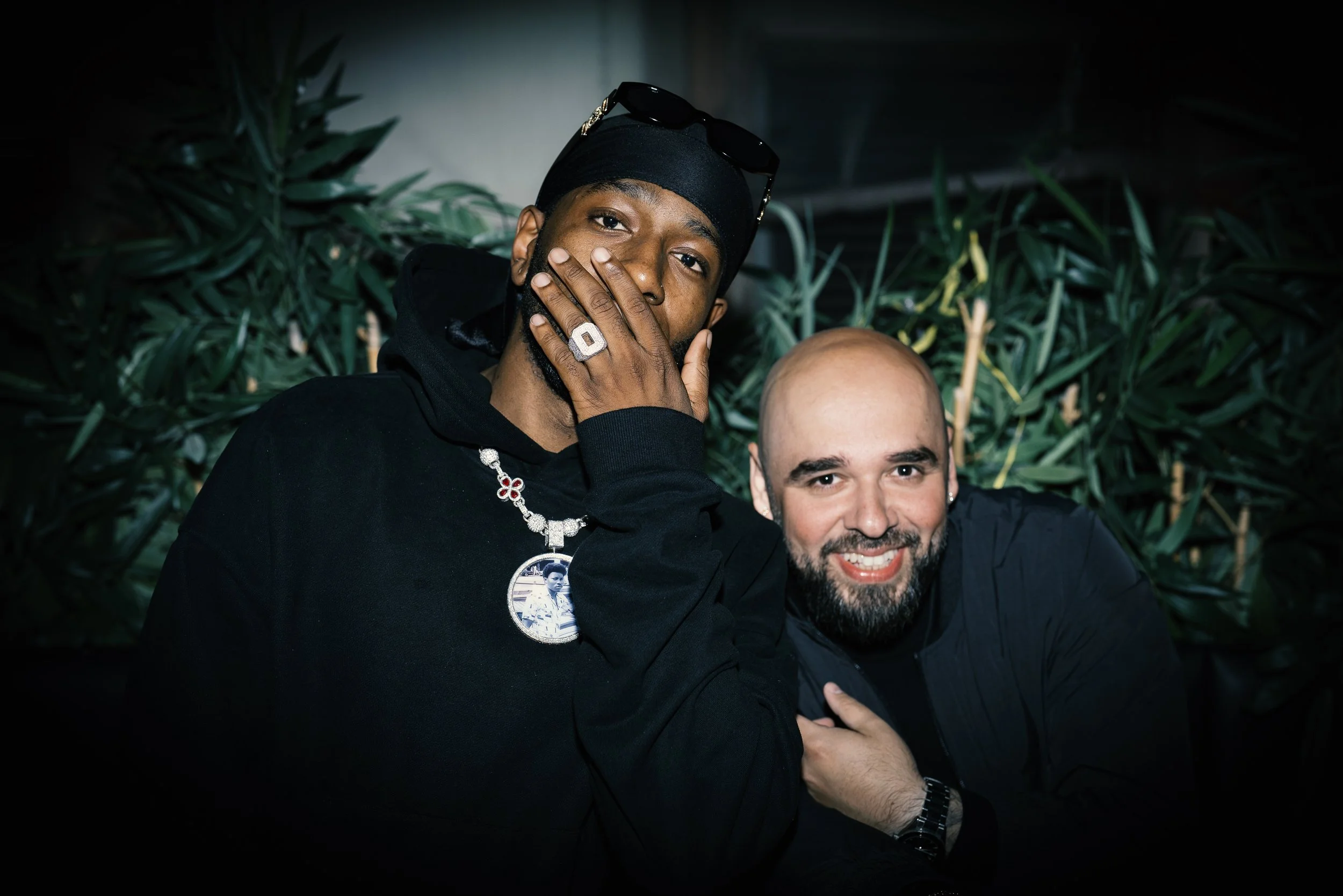 Two men posing for a photo, one with hand on face wearing jewelry and sunglasses on head, the other smiling and touching the shoulder. Background with green plants.