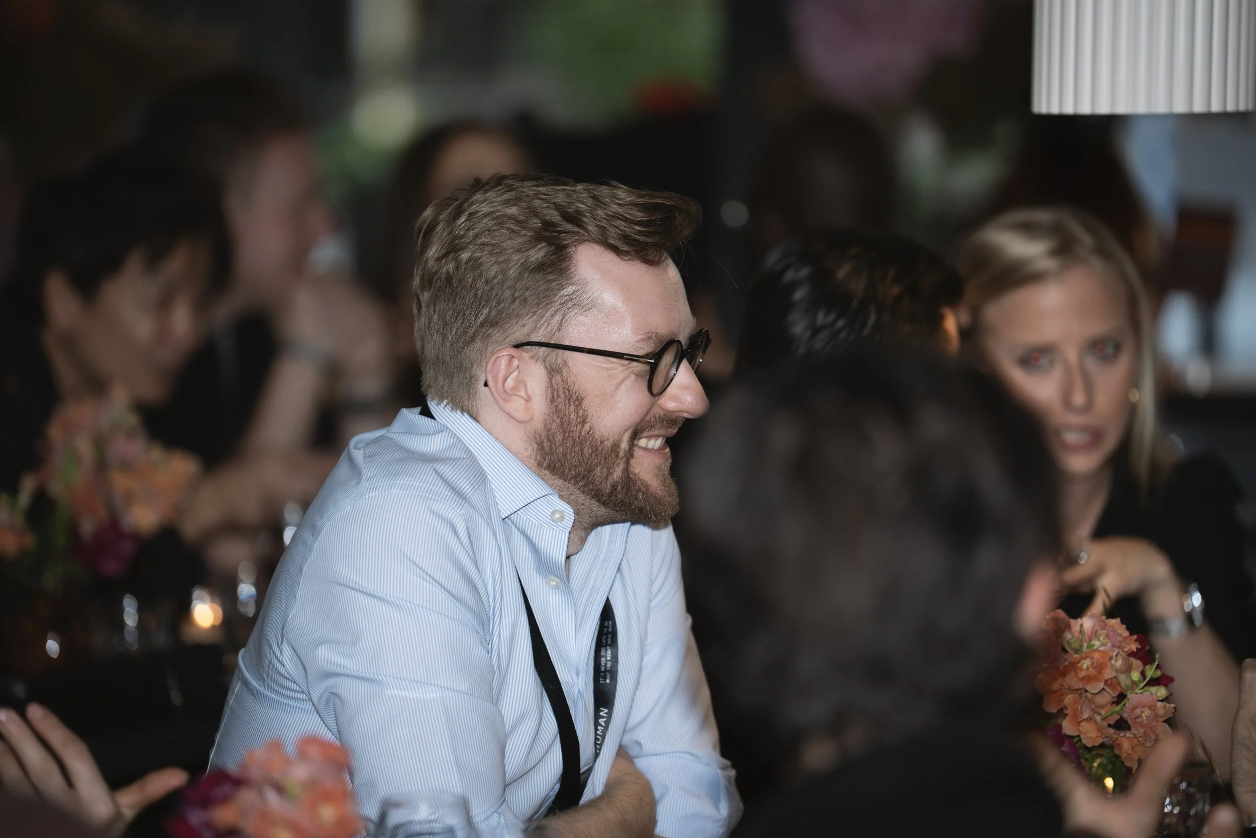 A group of people sitting at a table during a social event, with a man wearing glasses and a striped shirt smiling and engaging in conversation.