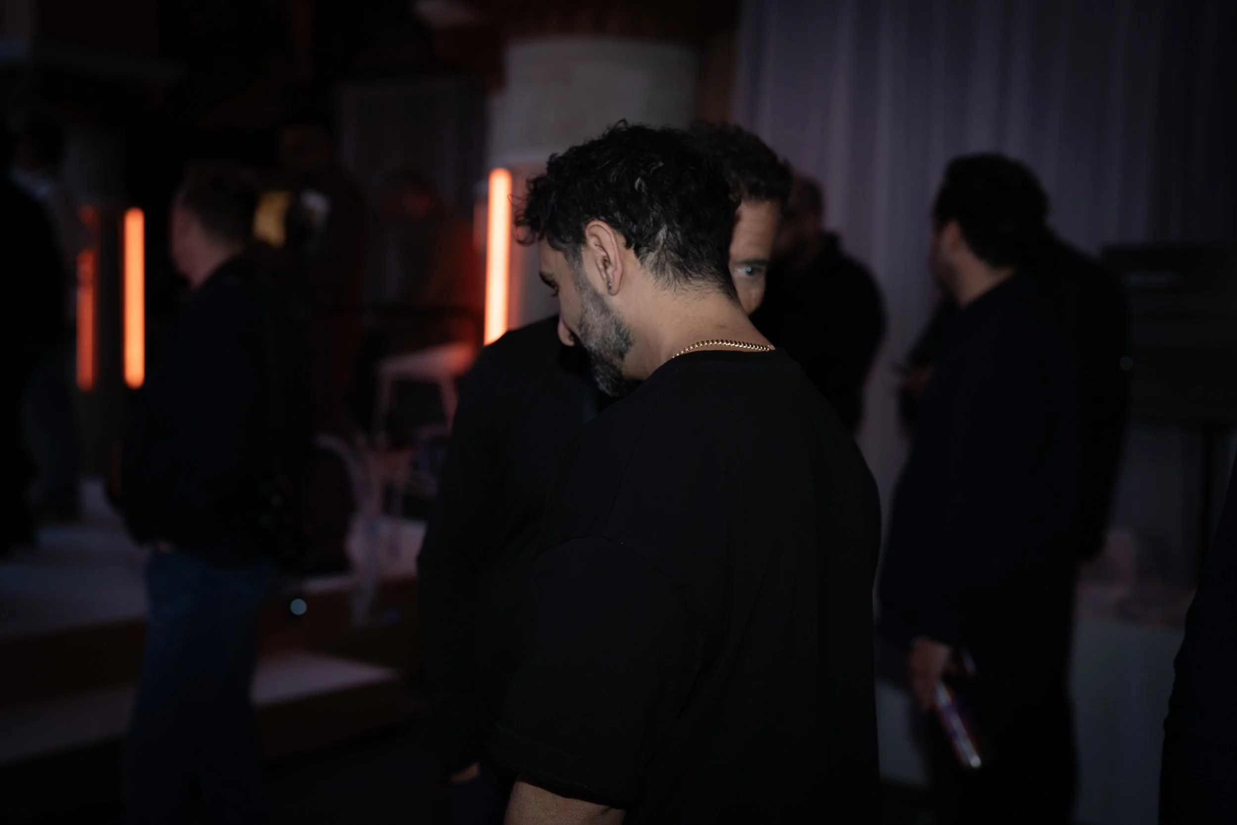 Man with dark curly hair and beard, wearing a black shirt and gold chain, standing with head slightly bowed in dimly lit room with red and purple lighting, surrounded by other people.